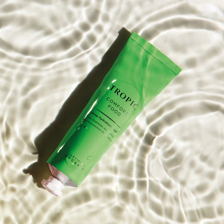 This image features a tube of Comfort Food, a deep hydration mask. The vibrant green packaging emphasises its nourishing and soothing properties. Key ingredients include wild Australian superberries and resurrection flower extract. The product is certified organic, and the rippling water effect in the background adds a refreshing, hydrating feel to the visual.